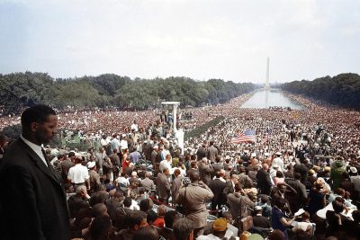 Photograph of Martin Luther King's "I have a dream" speech at Washington DC's Lincoln Memorial, with an enormous crowd gathered in front of him on stage, and some parts visible from behind the podium, looking towards the distant horizon where the famous obelisks and a large reflecting pool can be seen. In color. High resolution. In the style of ji hoches.