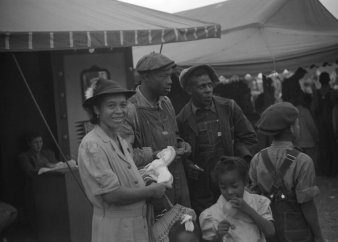 Black and white photo of African American people at the Texas state fair in front of an art deco tent, they have on work or overalls with flat caps and hats, one woman is holding her baby while another child chews gum. They look happy. The scene was captured in the style of Camera Kodak Brownie no 2a using black & white film.