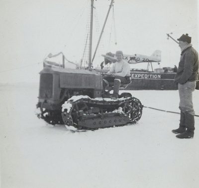 A black and white photo shows an old snow tractor on sea ice with two men in it, one holding onto its tracks while another man stands beside them smiling at the camera. In front, part of a yacht is visible which has "buy" written on top. Behind the car is a boat that says 'V compromisingatic'. The scene takes place during winter as shown by the snowy surroundings. One person wears a hat, jacket and boots. It seems to be taken from inside a ship looking out over the horizon in the style of Marcbye.