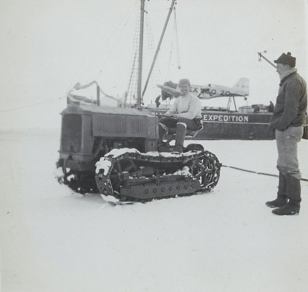 A black and white photo shows an old snow tractor on sea ice with two men in it, one holding onto its tracks while another man stands beside them smiling at the camera. In front, part of a yacht is visible which has “buy” written on top. Behind the car is a boat that says ‘V compromisingatic’. The scene takes place during winter as shown by the snowy surroundings. One person wears a hat, jacket and boots. It seems to be taken from inside a ship looking out over the horizon in the style of Marcbye.