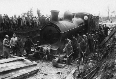 A black and white photograph shows the front side of an old steam locomotive. One person stands on it, with people around him as they work to sand down its wheels from debris after running at speed over small wooden sleepers. The photo was taken during World War I when French soldiers were building a train track near Stlandard for several days in winter. People were working hard wearing very simple uniforms. They all wore helmets or hats made of cloth. Some women were also present.