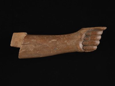 A carved wooden arm, with the hand pointing to one side and an outstretched finger on it. The entire arm is made of wood, showing signs of wear over time, with its edges slightly chipped or cracked due to use in ancient times. It's lying flat against a black background, emphasizing its simple shape and natural texture. This piece has been created as part of my series representing historical objects from different eras, capturing their unique characteristics and aesthetic appeal.