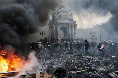 Photo of the large protest in Kyiv. Black smoke from burning trash and tires fills part of the frame. A group gathered around an old white baroque building with a black domed roof and golden accents. Black clouds overhead, black liquid on the ground near broken windows. A small figure stands at the far right wearing an orange helmet holding a green banner with a red symbol. Some people wear yellow caps over helmets or scarves. Many people wearing protective gear. A leader standing next to a pile of wood planks and a metal beam, in the style of [Banksy](https://goo.gl/search?artist%20Banksy).