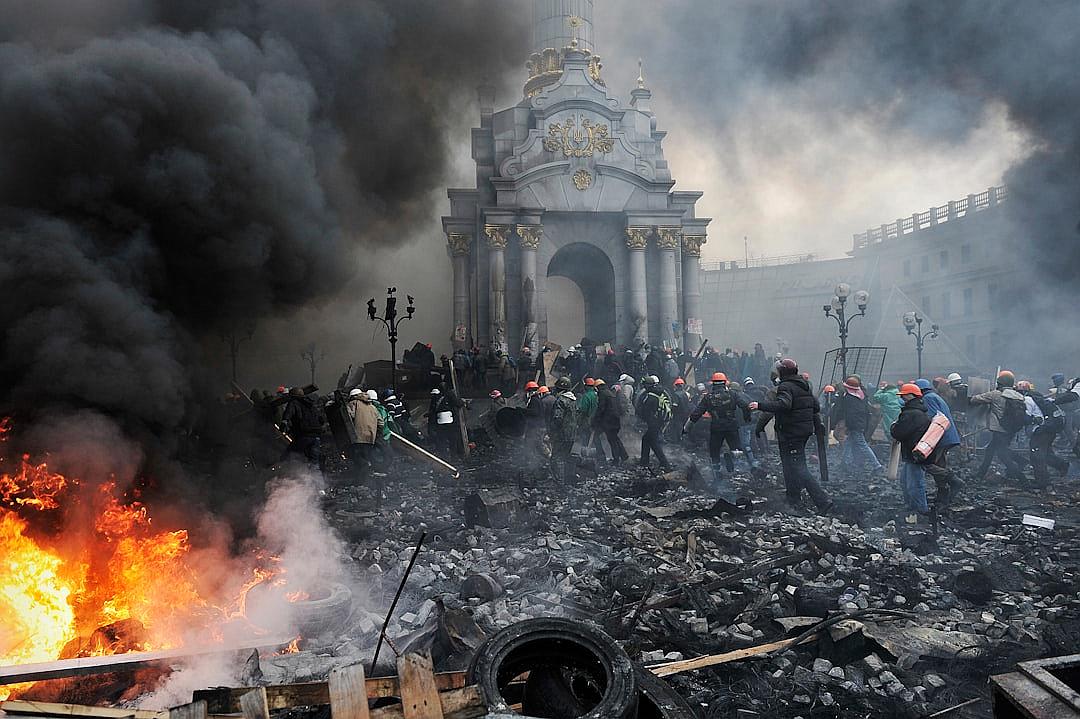 Photo of the large protest in Kyiv. Black smoke from burning trash and tires fills part of the frame. A group gathered around an old white baroque building with a black domed roof and golden accents. Black clouds overhead, black liquid on the ground near broken windows. A small figure stands at the far right wearing an orange helmet holding a green banner with a red symbol. Some people wear yellow caps over helmets or scarves. Many people wearing protective gear. A leader standing next to a pile of wood planks and a metal beam, in the style of [Banksy](https://goo.gl/search?artist%20Banksy).