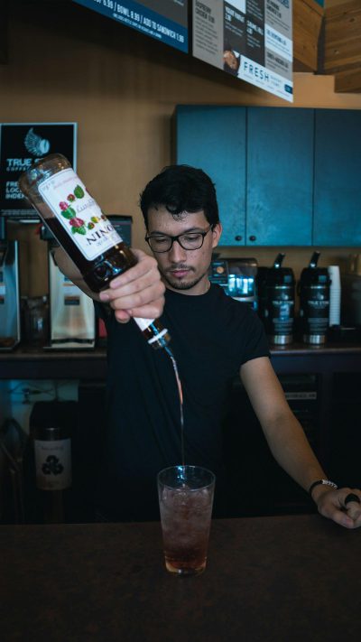 Photo of a professional mixologist pouring liquid from one bottle into another in front of him wearing a black t-shirt and glasses, behind him is a modern coffee shop bar with blue accents, a glass in hand for a full product shot, product photography, the bottle label says "by FAlan Bredman"