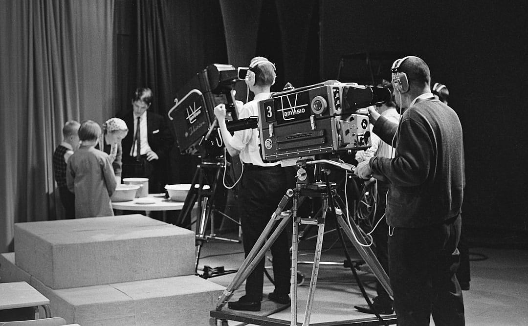 A black and white photo of the camera crew filming the presenters on stage in front of an audience at a TV studio, in the style of 60s style, with a film grain effect, featuring an old tv set.