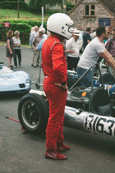 A candid photo of an F2 race car driver wearing red racing suit and white helmet, standing next to his blue vintage formula one in the pit area atwell green village festival with people around him and other cars parked on street,