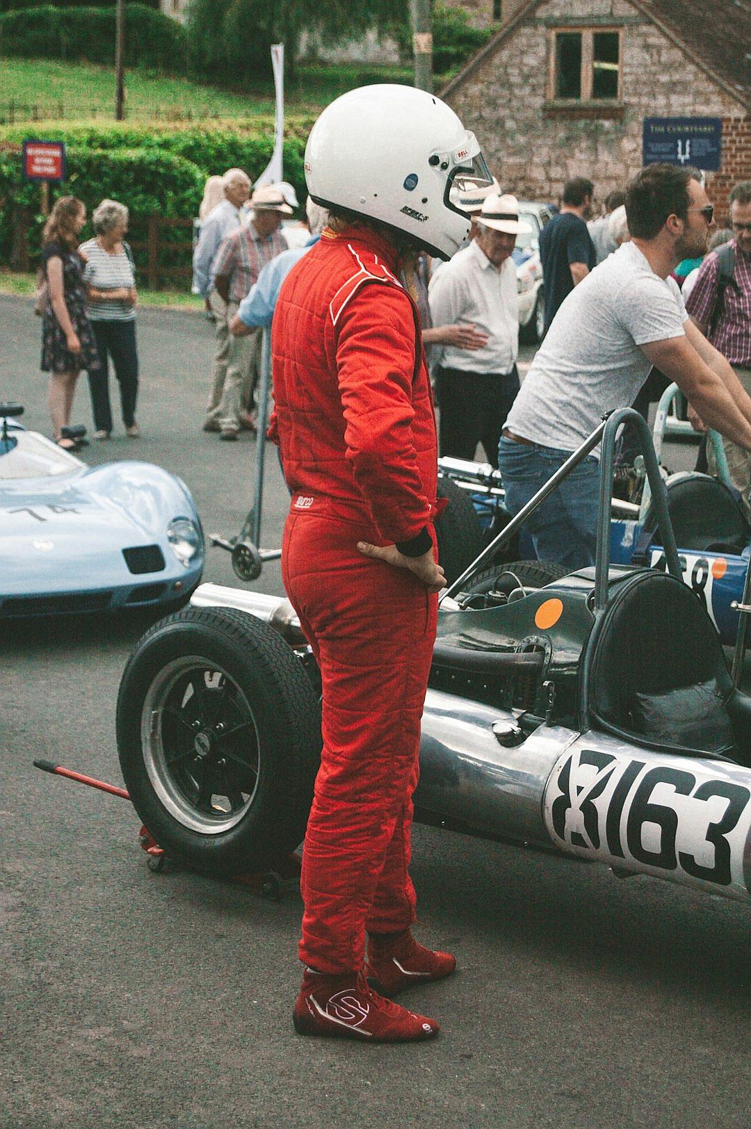 A candid photo of an F2 race car driver wearing red racing suit and white helmet, standing next to his blue vintage formula one in the pit area atwell green village festival with people around him and other cars parked on street,