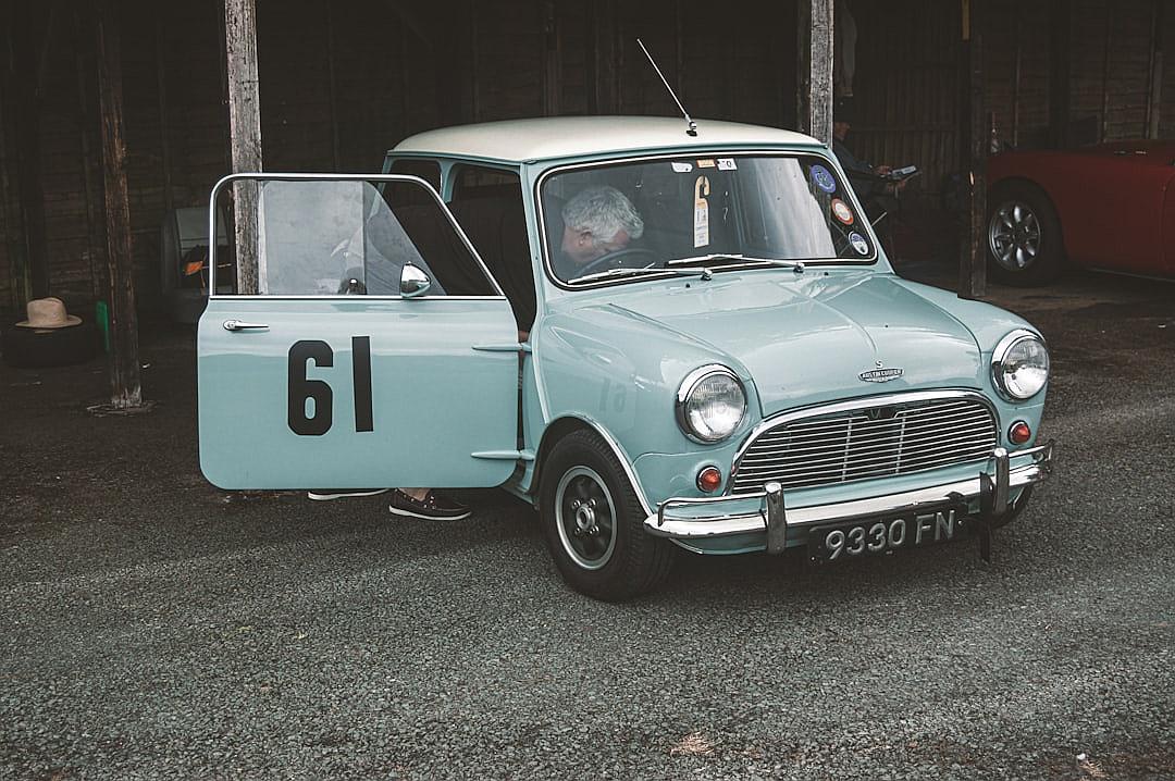 A light blue vintage mini with black numbers on the doors, racing in an old English garage. A middle aged man sitting inside it in the style of vintage photography and in a retro style.