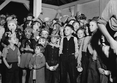 A group of children in costumes stand at the entrance to an open-air dance hall, surrounded by adult men and women who all wear masks, in the style of black-and-white photography from the 1950s American drama era.