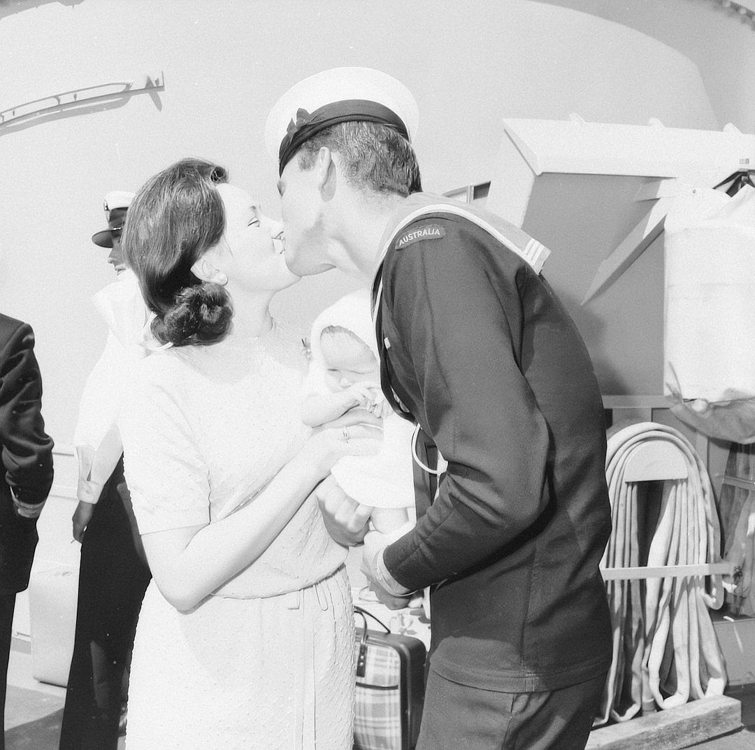 A black and white photo of an American sailor in uniform, on the deck of a ship holding his newborn baby while he kisses her cheek with their mother standing beside them smiling at both. They all wear white dresses. The photo is in the style of [Ansel Adams](https://goo.gl/search?artist%20Ansel%20Adams).