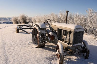 An old tractor with snow on it is parked in the middle of an empty field covered in white, thick ice and frost. The background features low shrubs covered in fresh rime, creating a picturesque winter landscape. This scene captures intricate details and textures, emphasizing both nature's beauty and vintage machinery. Photography captured the scene using a wideangle lens to capture more of the surroundings, in the style of a vintage photograph.