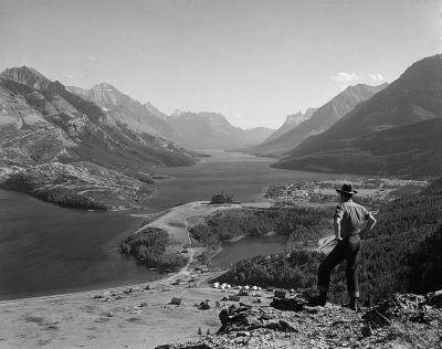 A black and white photograph of British explorer John semenchuk standing on the edge overlooking Ancient Lake in grocery national park, a view from above showing a valley with a town at its base. Taken in the style of [Robert Capa](https://goo.gl/search?artist%20Robert%20Capa) using an old camera from the 1920s. The man is wearing jeans, a shirt and cowboy hat, he's leaning against his left shoulder looking over the water. In the background, mountains and forests surround the lake. High contrast. A vintage feel. Shot on Kodak film.