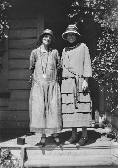 Two women in long dresses and hats standing on the front porch of their house, in a black and white photograph from the early 20th century, posing for a photo with smiles, in a full body shot, in summer , with neutral expressions, photographed with a 35mm lens at an f/8 aperture with natural lighting on a sunny day.