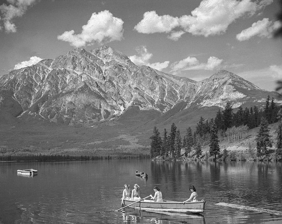 A black and white photograph of people in canoes on definition lake with rocky mountains behind, trees along the shore, some women wearing vintage swimsuits. A few clouds scattered across sky. Taken by Hasselblad camera.