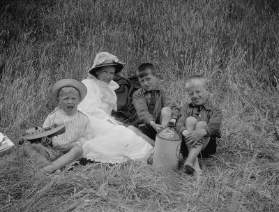 An old black and white photo of happy kids playing with a milk carton in the grass, a mother sitting next to them wearing a long dress and hat.