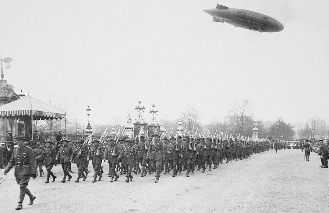 Black and white photograph of the WWI French heavy armored guard, marching in line through Champs Elysées Paris with an airship floating overhead. High resolution photograph with photo quality, in the style of an early 20th century photographer.