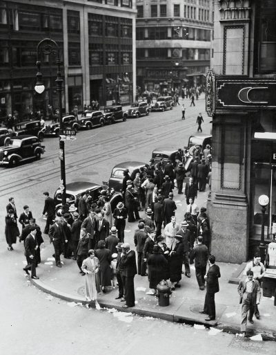 A black and white photograph of the streets outside an art deco style bank in Chicago, with people standing on street corners waiting to get into it during downtown business hours. The photo is taken from above looking down at them, with cars driving by. It's 20s era America.