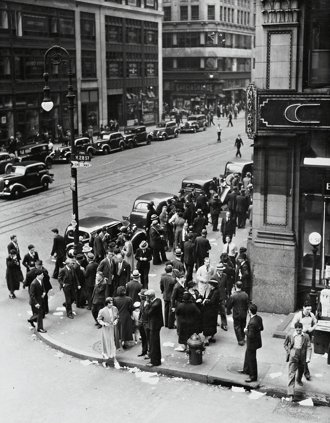 A black and white photograph of the streets outside an art deco style bank in Chicago, with people standing on street corners waiting to get into it during downtown business hours. The photo is taken from above looking down at them, with cars driving by. It’s 20s era America.