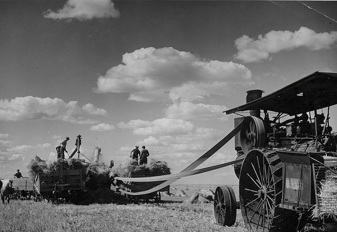 A black and white photo of an old-fashioned harvester in the field, with people working on it to harvest crops. The sky is clear blue with fluffy clouds, adding depth to the scene. In front of them stands another machine that has ribboned straw from grain and points towards workers who stand nearby. This photo was taken in the style of [Man Ray](https://goo.gl/search?artist%20Man%20Ray) during his time at Madoxins Beach School.