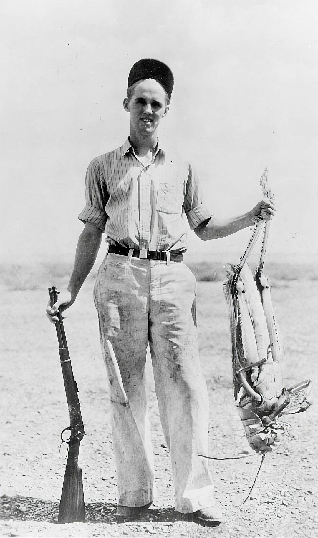 A young man in white trousers, black shoes and with his hair combed to the side stands on an open field holding rifle at waist height. He smiles while posing for photo with dead fish hanging from both hands. The background is desert landscape. Black & White Photo. 20th century style. High resolution