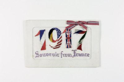 A white rectangular embroidered patch with the number "207" in large letters, adorned in the style of flags of France and Great Britain. The text is written on it as well "Souvenir from france". A ribbon hangs delicately around the corner of that paper sticker. White background.