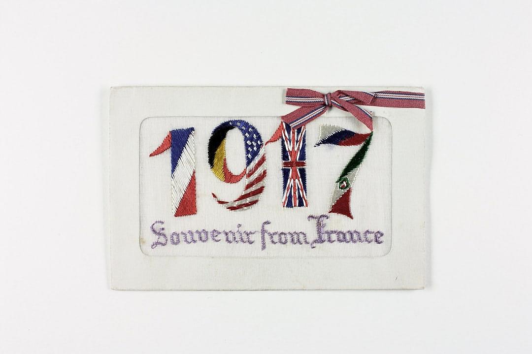 A white rectangular embroidered patch with the number “207” in large letters, adorned in the style of flags of France and Great Britain. The text is written on it as well “Souvenir from france”. A ribbon hangs delicately around the corner of that paper sticker. White background.