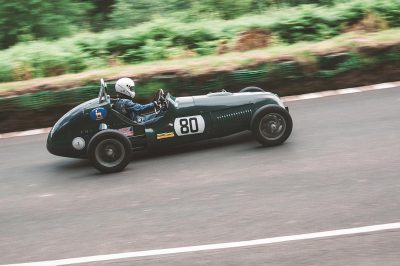 Vintage racing car with the number "80" on the side, a driver in a helmet and blue suit driving fast down an English road, a green forest background, a motion blur effect, a side view, a wide angle shot, a vintage color grading, an f/2 lens, a Leica M3 camera, a grainy film texture.