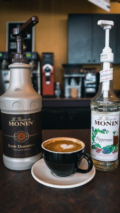 A photo of the "Le Texte de Monin" dark chocolate and Peppermint colored cream coffee bottle next to an espresso cup on top of cafe counter, with milk frother machine and bottles of white sugar in the background. The scene is captured from above in the style of Canon EOS R5 mirrorless camera using a wideangle lens.