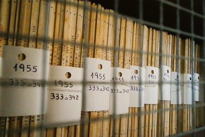 A closeup of the bottom side of white tags with numbers "325" and text on them, hanging from an iron wire shelf filled with filestacks labeled in black ink as if they were documents or backgrounds, each either covered by several rows of very thick white paper. The scene is illuminated by natural light that highlights details like the soft texture of the beige wood grain. In the background, outoffocus lines lead to more equipment behind it, adding depth and dimension to the composition.