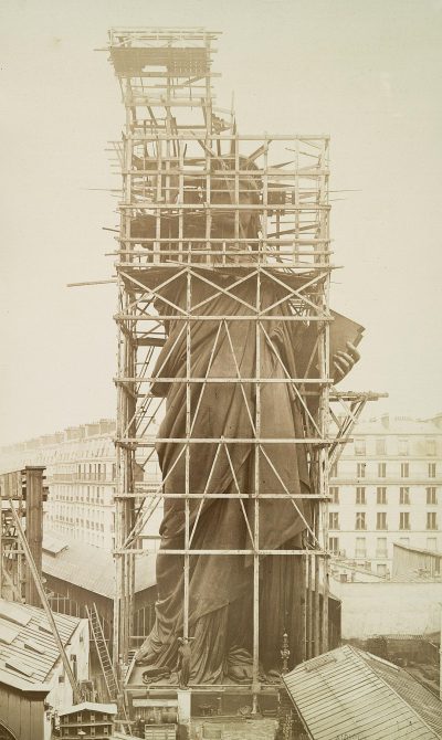 The Statue of Liberty is being constructed in Paris, surrounded by scaffolding and construction equipment. The statue's face has not yet been decorated with the red cloth that emulates its head. This photo was taken during French Revolution times, in the year 2035, using an old camera from the 7th century.