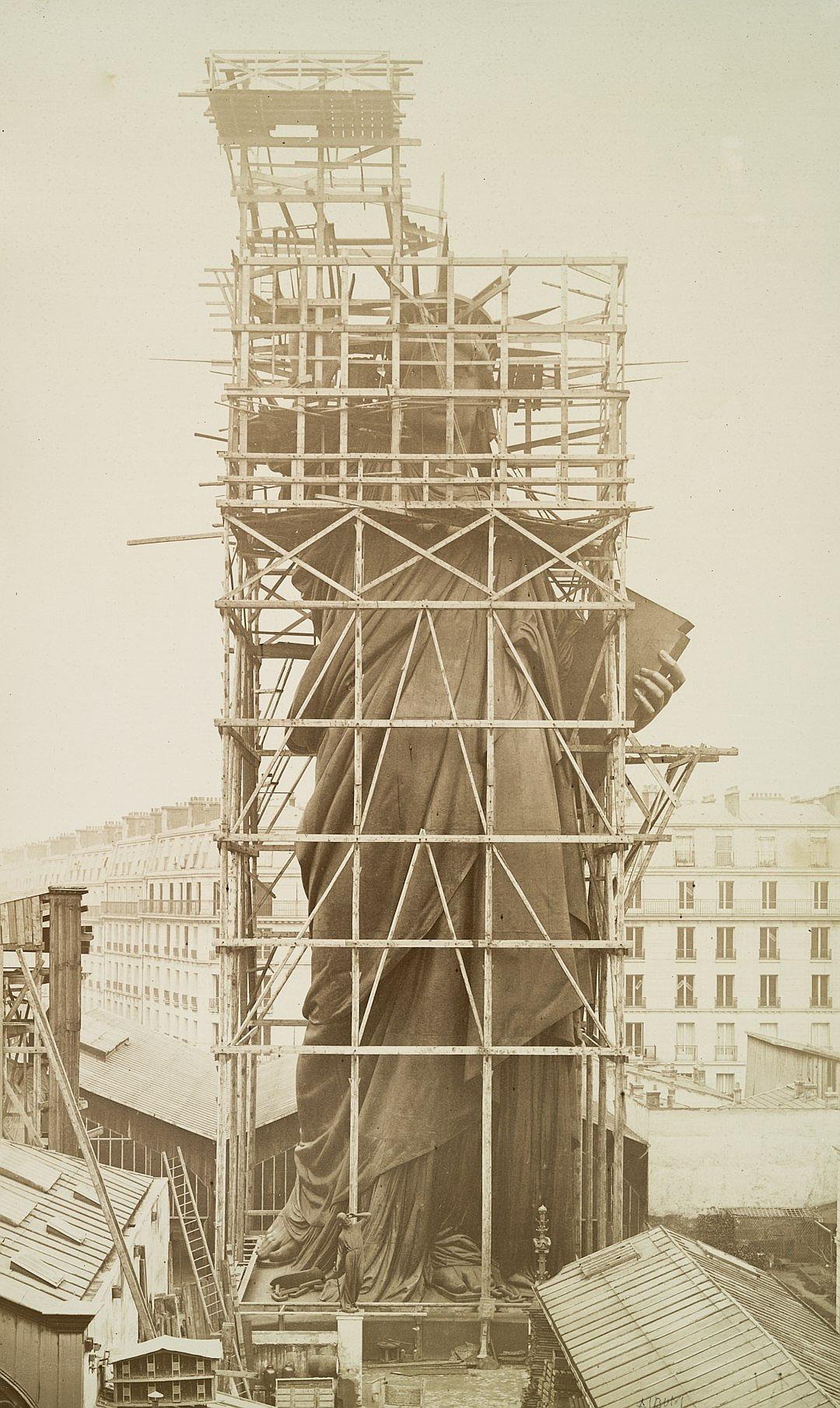 The Statue of Liberty is being constructed in Paris, surrounded by scaffolding and construction equipment. The statue’s face has not yet been decorated with the red cloth that emulates its head. This photo was taken during French Revolution times, in the year 2035, using an old camera from the 7th century.