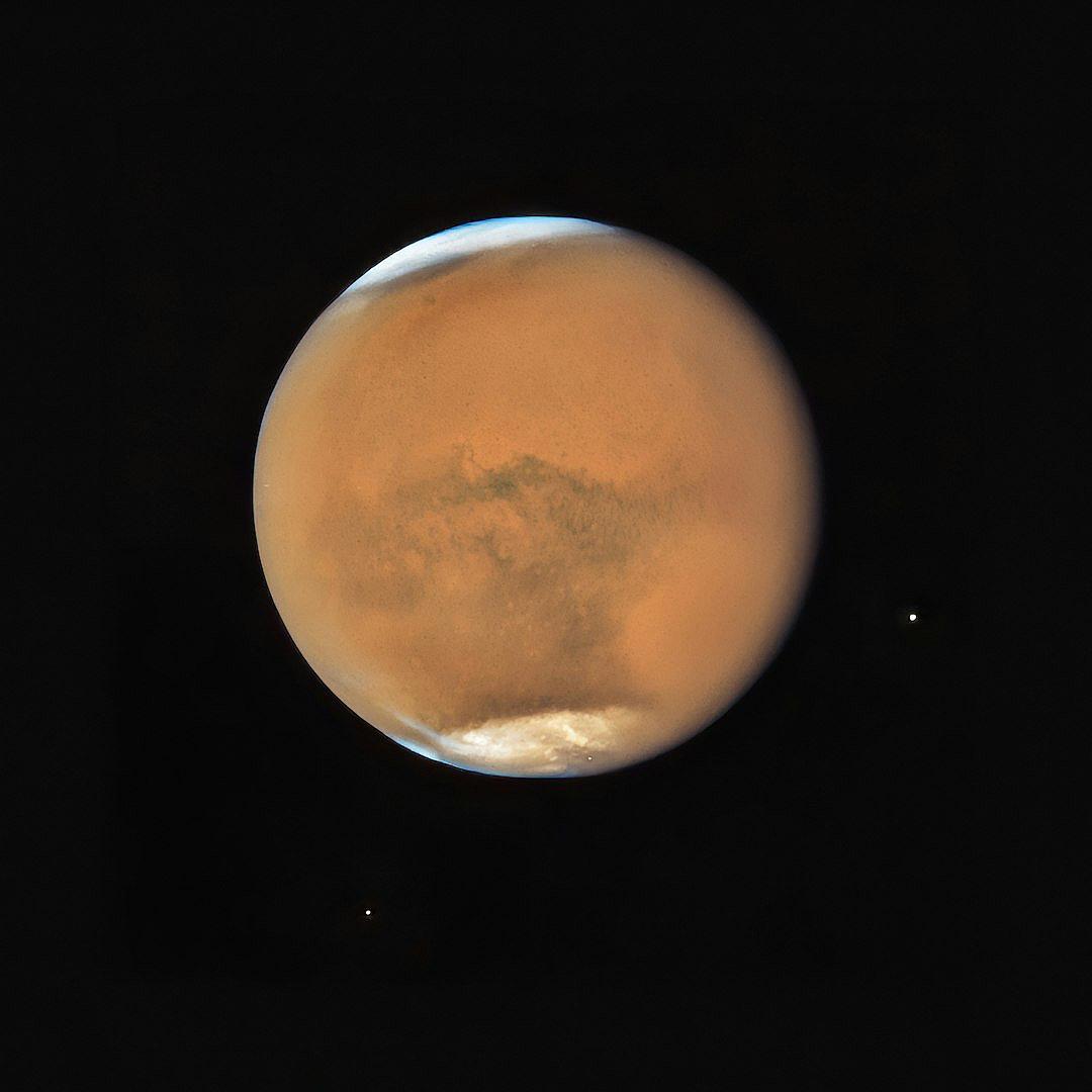 A photograph of the planet Mars taken by the Hubble Space Telescope, with the planet centered in the frame against a black background. The atmosphere on Mars looks thick and red, with a small moon just above its horizon. The moon appears to be a blueish white color, in the style of Hubble Space Telescope.