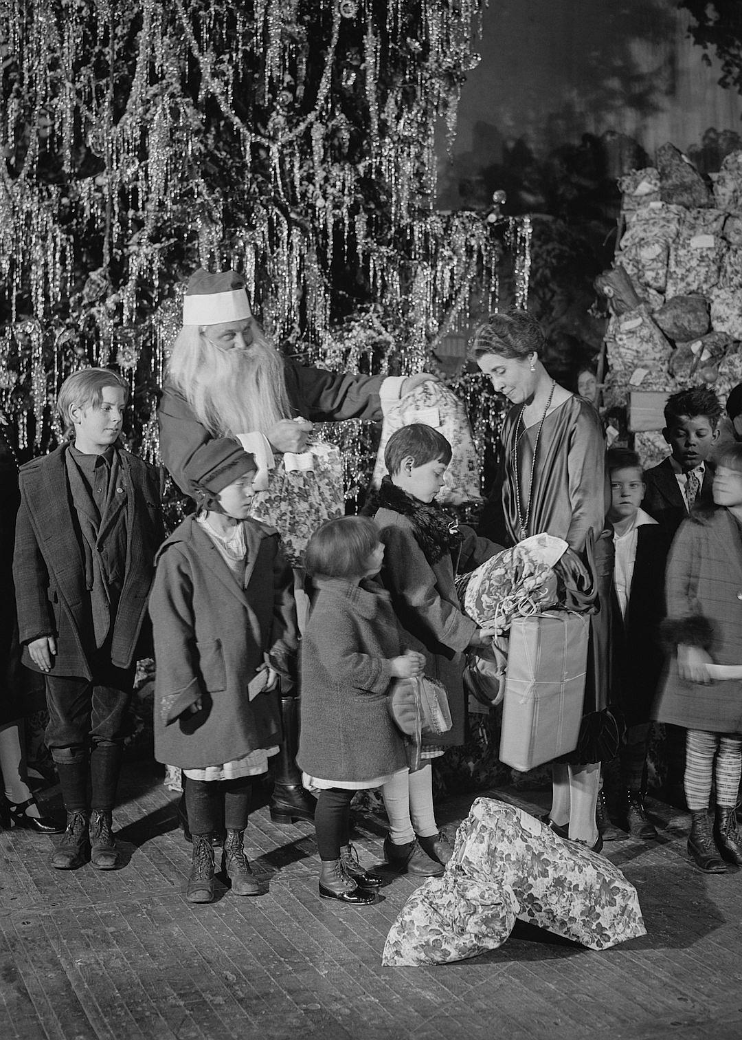 old black and white photo of santa claus giving gifts to children, victorian style, christmas tree in the background, people standing around, christmas decorations on wall, flowers and garlands decoration, 20s photography