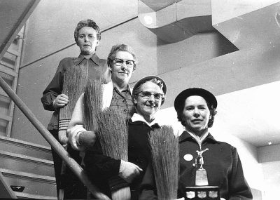 A black and white photo of four women holding long wooden brushes, standing on the staircase in an office building. They all wear work with baseball caps over their hair. One woman is smiling while another has her head down. The other two have one hand resting against each shoulder and hold what appears to be some type of trophy or award. In front them there's a large piece of paper hanging from above that could be used for taking notes or drawing graphics. On top of it we can see three arrows pointing up. It seems like they were celebrating something because one holds a brush as if showing off its long handle. There may also be