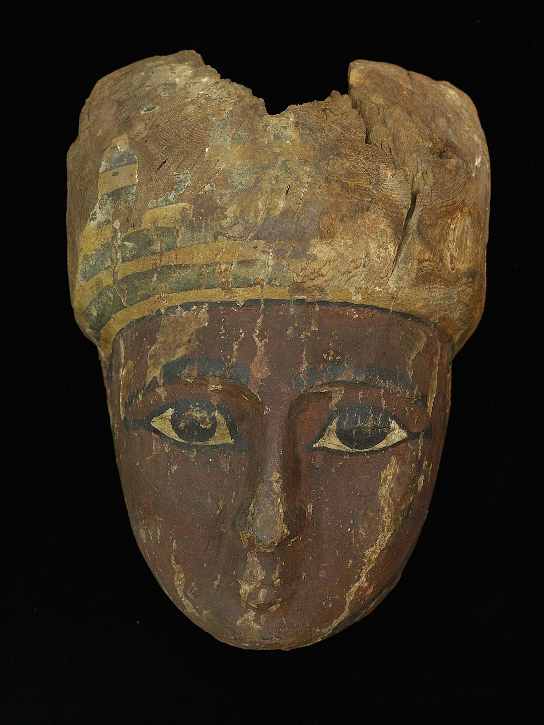 An ancient Egyptian woman’s head, painted on wood and worn by time with no hair or eyebrows, carved out of an oval shaped piece of stone in the style of Hategic. A oakwood mask with gold edging around the eyes, painted face, small horns, black background.