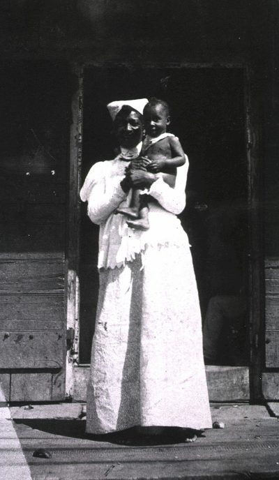 African American woman wearing a white dress and nurse cap standing on the porch of an old wooden house holding up her baby in a black and white photograph, in the style of an early 20th century photographer.