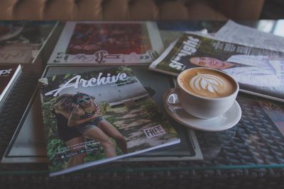 A magazine on the table with coffee and other magazines, titled "staggering", features a photo of a woman in sports  wearing high heels on the cover. A white cup of cappuccino sits next to it, with title text saying "orda", and text below advertising 'free archiving'. The magazine lays flat on the table with the coffee cup beside it.