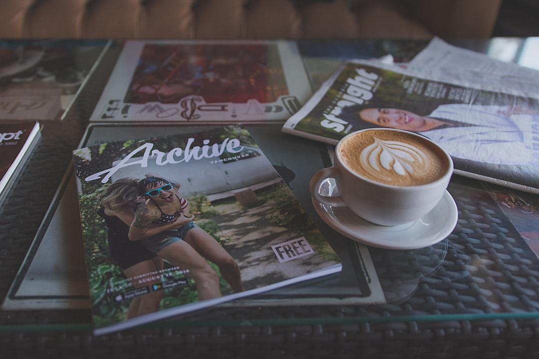 A magazine on the table with coffee and other magazines, titled “staggering”, features a photo of a woman in sports  wearing high heels on the cover. A white cup of cappuccino sits next to it, with title text saying “orda”, and text below advertising ‘free archiving’. The magazine lays flat on the table with the coffee cup beside it.