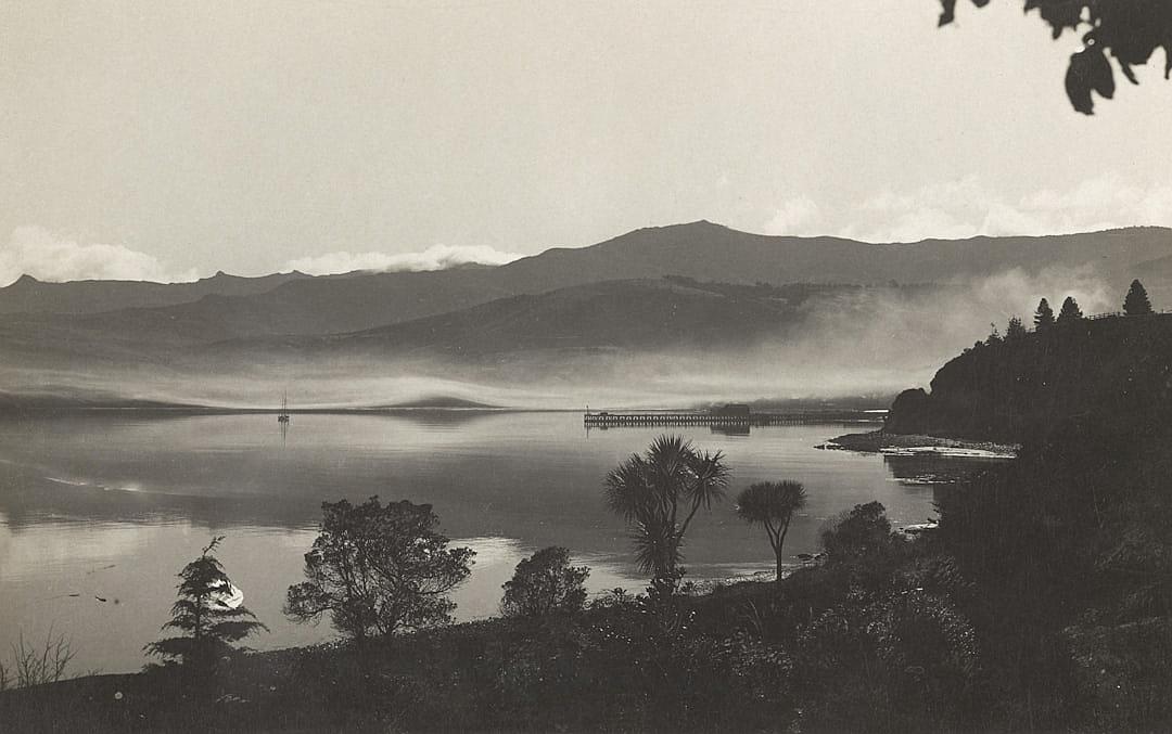 photograph of lake bank with mountains in the background, black and white photo in the style of Paul caponigdi, New Zealand landscape, 20th century photography, view from hilltop overlooking the water with pier extending into it, trees and plants on hillsides, distant mountain range in hazy mist,