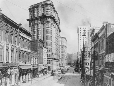 vintage black and white photo of the corner building on the right side, in downtown Charleston old tall buildings with large windows, streetcar track overhead, people walking along busy city streets in the style of Controllers' League of America.