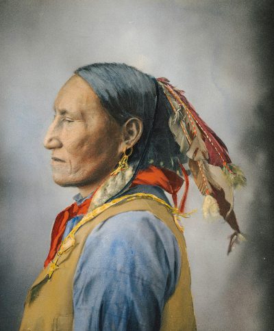 A vintage color hand tinted film grain portrait of an old native american man with long hair and wearing traditional , profile view, side angle shot