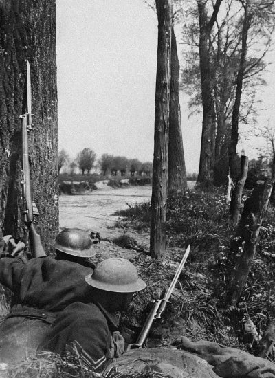 In the woods of French Flanders, in winter with no leaves on trees and a field visible behind them, two British soldiers from World War One sit against tree trunks while holding their rifles up to cover themselves as they watch for Germans behind the bushes. They wear World War One helmets but without gas masks. The photo is taken in the style of William Häenick. It's a black and white photo taken during daytime.