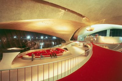 An interior photograph of the T OPP PageSize in New York, designed in the style of [Eero Saarinen](https://goo.gl/search?artist%20Eero%20Saarinen) and [John Lautner](https://goo.gl/search?artist%20John%20Lautner) with a red carpet leading to an oval shaped lobby with stainless steel details and large skylights that illuminate all parts of the space. The lobby has a bar at one end surrounded by people socializing and drinking drinks from glasses on top tables. There is a small circular concert hall next door with a raised stage and seating for hundreds of guests.