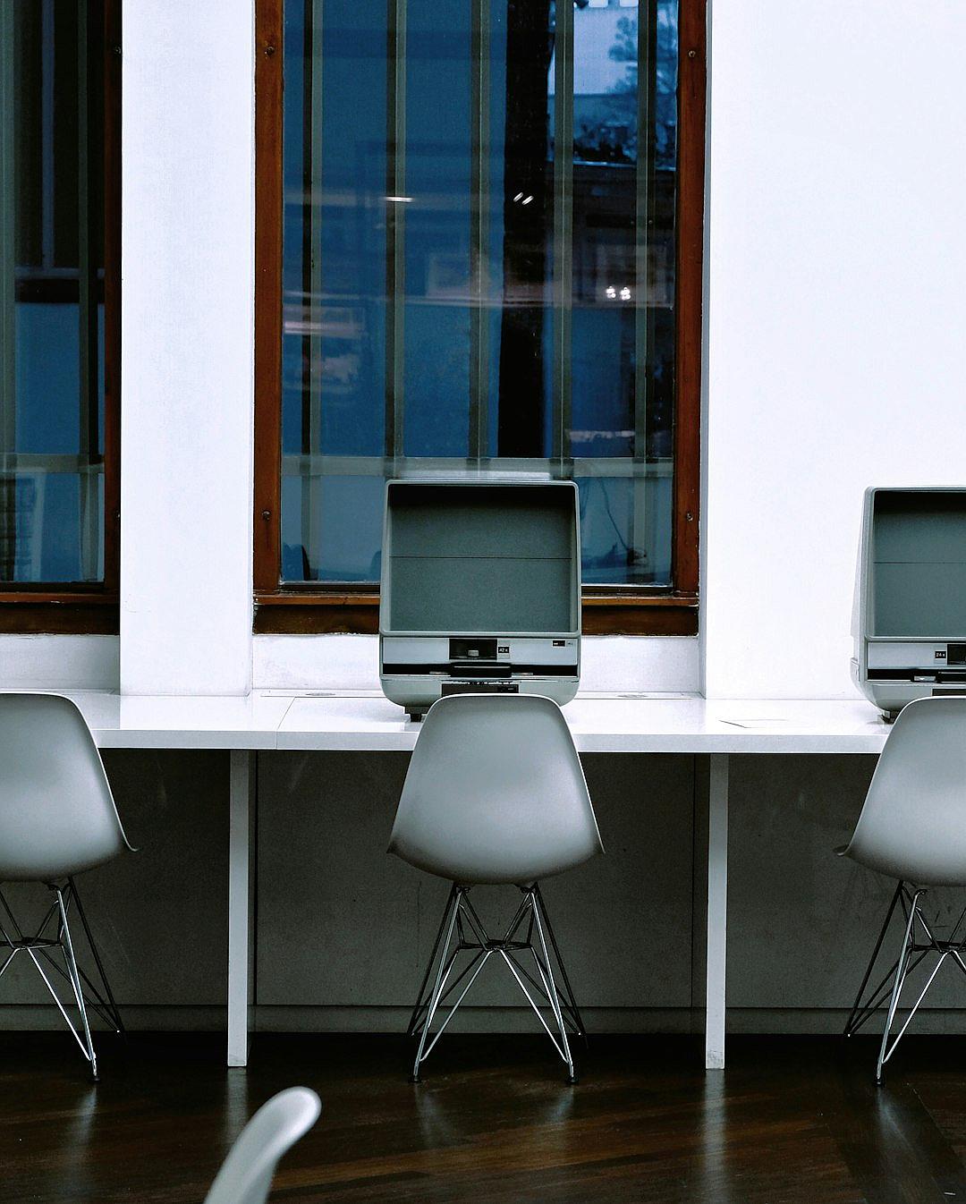 A row of computer desks with gray chairs, white walls and windows in the background. The computers have oldschool monitors on them. Shot from behind the desk. Photo taken by [Mario Testino](https://goo.gl/search?artist%20Mario%20Testino) using a Canon EOS camera.