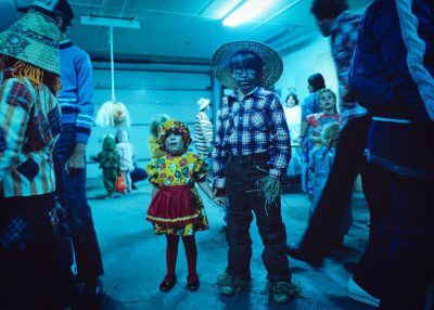 color photo of two children dressed in blue and yellow with black faces, they dance at the end of an adult party wearing costumes made by their parents, they stand next to each other on one side of them is another child who wears colorful while others look around him in horror, inside a warehouse illuminated by neon lights, kodak film photography