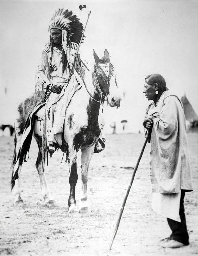 black and white photo of an Indian chief on horseback, with his spear in hand talking to someone who is standing next to him wearing long robes and walking stick. They both look at each other. In the background you can see tents behind them, they appear small in comparison to their huge horses which have black hoods over their heads, which cover all three ears, and also make it hard to tell if he has hair or not. It's a clean, clear sky with no clouds.