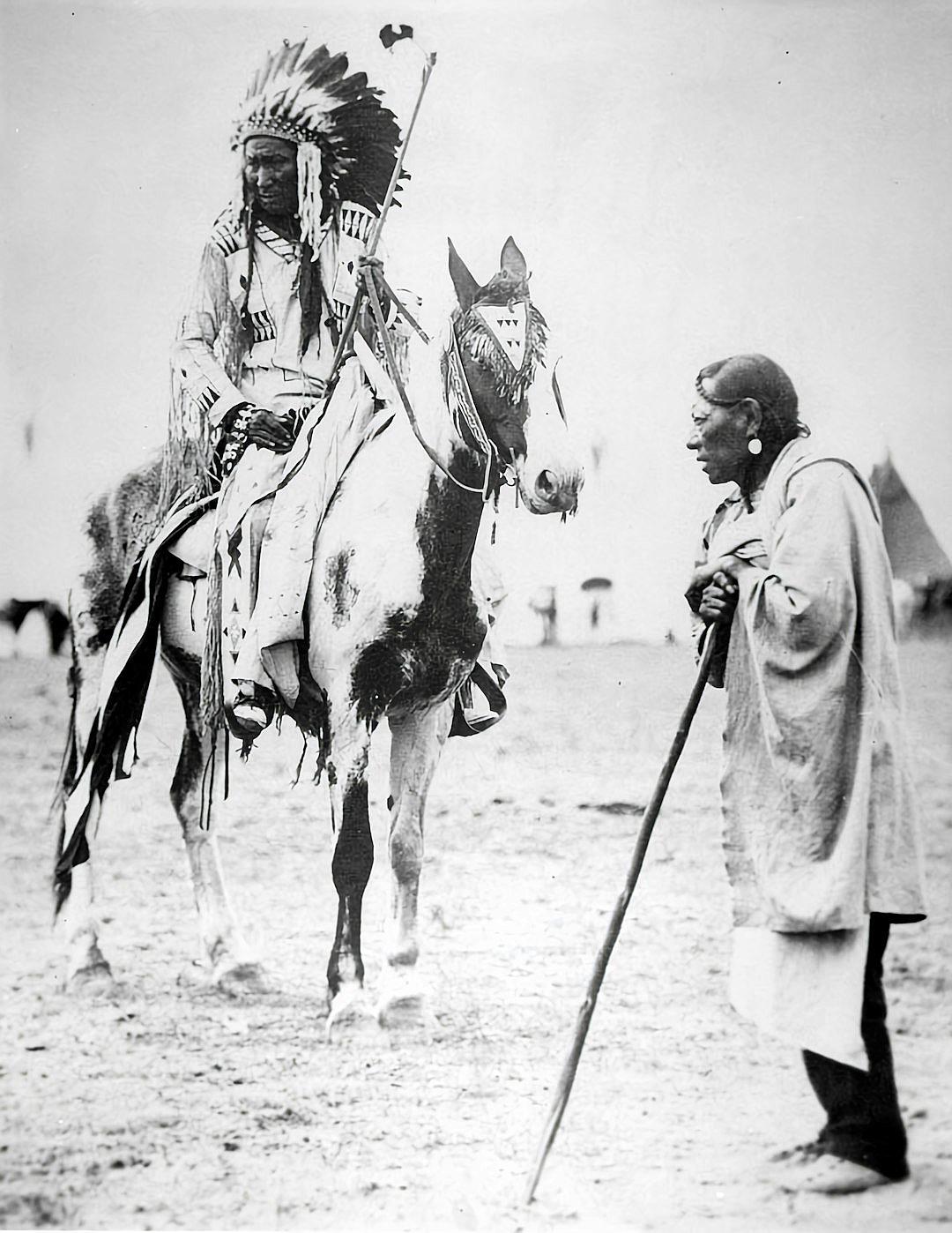 black and white photo of an Indian chief on horseback, with his spear in hand talking to someone who is standing next to him wearing long robes and walking stick. They both look at each other. In the background you can see tents behind them, they appear small in comparison to their huge horses which have black hoods over their heads, which cover all three ears, and also make it hard to tell if he has hair or not. It’s a clean, clear sky with no clouds.