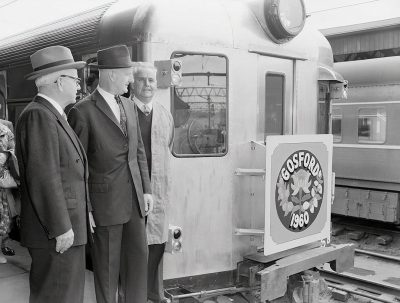 Black and white photo of three men in suits standing next to the door on an old train, with "Bos쇠 MD" written inside one window. The man is smiling at someone off camera while looking out from behind him through the open doors of his carriage onto platform, where people stand around waiting for their trains. On side wall of car there's large logo depicting maple leaf and letters 'teenth Mountain'. There's also sign that says 'flatMap Dagger