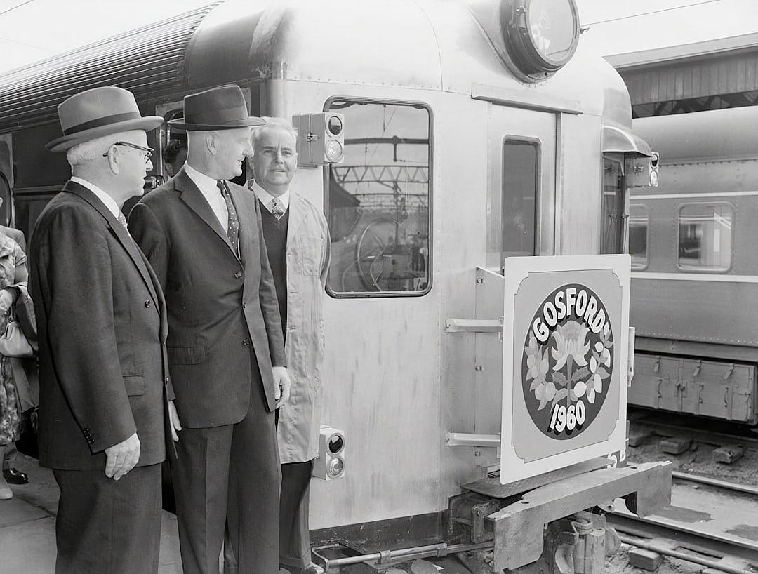 Black and white photo of three men in suits standing next to the door on an old train, with “Bos쇠 MD” written inside one window. The man is smiling at someone off camera while looking out from behind him through the open doors of his carriage onto platform, where people stand around waiting for their trains. On side wall of car there’s large logo depicting maple leaf and letters ‘teenth Mountain’. There’s also sign that says ‘flatMap Dagger